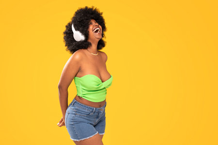 Adorable african american woman with white headphones laughing out loud, dancing, yellow vibrant studio background. Real people emotions. A lot of copy space.の写真素材
