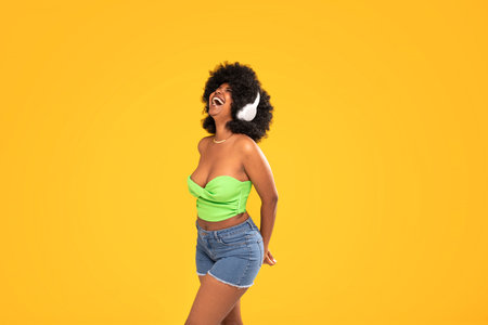 Adorable african american woman with white headphones laughing out loud, dancing, yellow vibrant studio background. Real people emotions. A lot of copy space.の写真素材