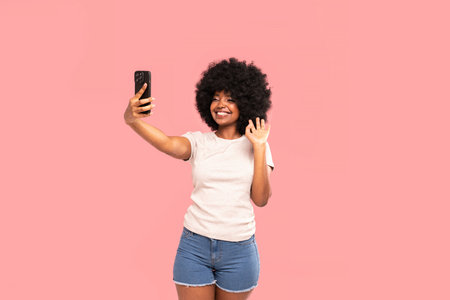 Young African American woman  with afro hairstyle holding phone and smiling, doing selfie or talking with friend online. A lot of copy space. Pink pastel studio background. Happy student.の写真素材