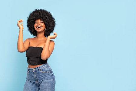 Portrait of cheerful young African American female model in black top and jeans, with afro hairstyle, smiling to the camera. Real people emotions. A lot of copy space. Blue pastel studio background.の写真素材