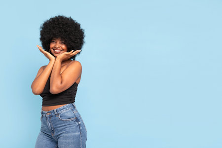 Smiling young girl with afro hairstyle and posing in black top and jeans, looking at the camera. Happy woman on blue pastel studio background. A lot of copy space.の写真素材