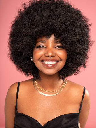 Close up studio portrait of happy girl with big smile and afro hairstyle. Smiling dark skinned beautiful woman looking at the camera. Positive female wearing gold jewelry, necklace. Pink pastel background.の写真素材