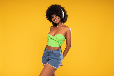 Energetic, happy, young woman with afro hairstyle dancing, laughing, listening music with wireless headphones. Yellow studio background. Copy space.の写真素材