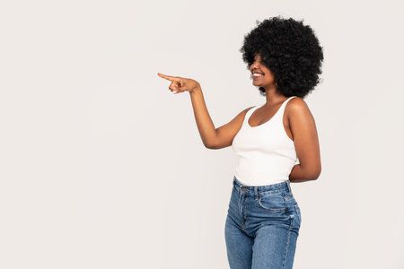 Happy african-american girl with afro hairstyle and big smile pointing on the empty studio background. Young woman in white top and jeans. Sale concept.の写真素材