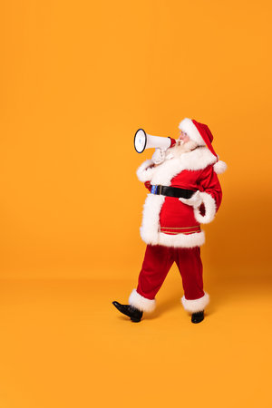 Christmas is coming! Real Santa Claus shouting using megaphone over yellow studio background. Xmas sale, discount concept. Copy space. Full length photo.の写真素材
