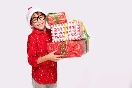 Cheerful funny boy in red Santa hat holding Christmas gifts on white studio background, smiling to the camera. Xmas time.の写真素材
