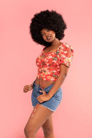 Photo of charming african american fashionable female in trendy jeans shorts posing over pastel pink studio background. Girl looking at the camera with delicate smile. Beauty woman with afro hairstyle.の写真素材