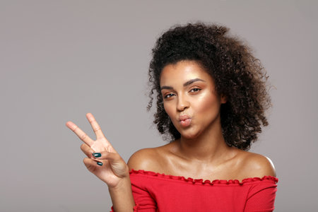 Playful positive curly haired woman send a kiss, shows a happy peace gesture. Real people emotions concept. Studio shot. Beauty portrait.の写真素材