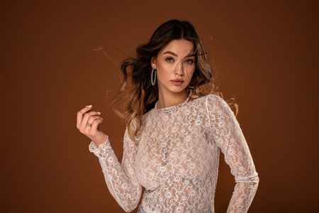 Beautiful young natural woman with delicate makeup in romantic lace dress and fashionable jewelry posing on studio background. Bride to be with long wavy hair. A lot of copy space on the brown studio background.の写真素材