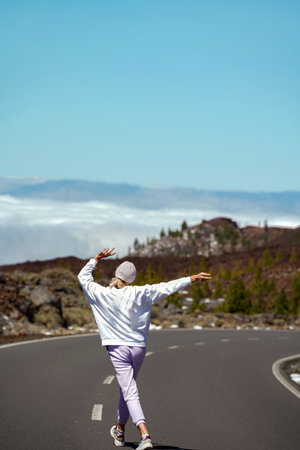 Happy woman running on the beautiful mountain road, looking forward with arms raised, view from the backside. Lifestyle travel concept. Tourist. Wanderlust.の写真素材