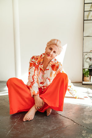 Elegant, fashionable mature blonde woman relaxing, sitting on the floor, looking at the camera with delicate smile.の写真素材