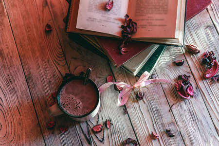 Mug of hot chocolate, dry fragrant rose petals, and old books on wooden table. Vintage filter style, top view.の写真素材