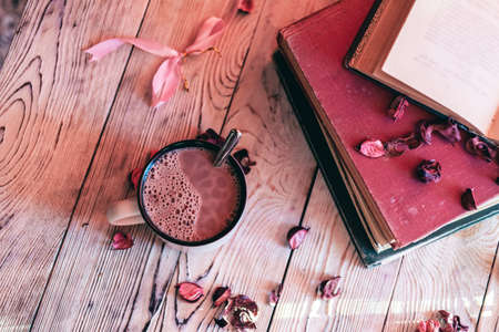 Cup of hot chocolate, dry fragrant rose petals, and old books on wooden table. vintage filter styleの写真素材