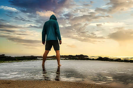 Man in blue sweatshirt with feet in the ocean before sunset