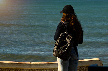 Woman with a cap and backpack looking out to sea from the seafrontの写真素材