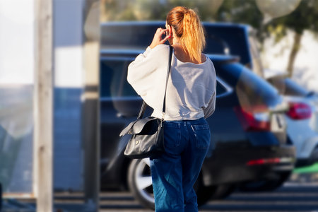 Young red-haired woman in a white shirt and jeans with a black handbag is standing on the background of a car.の写真素材