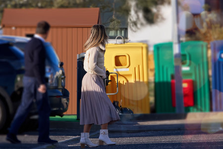 Young woman with long blonde hair, dressed in a long skirt and white blouse, stands near the yellow trash can on the street.の写真素材