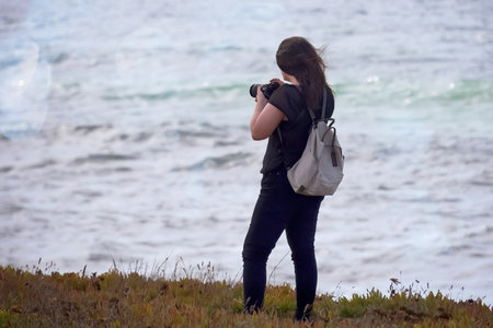A young female photographer taking a picture of the ocean on a sunny day.