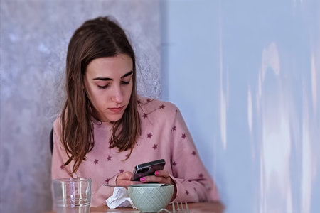Young woman in pajamas sitting at the table and using smartphoneの写真素材