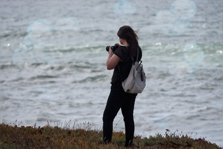 Young female photographer taking photos of the sea. She is wearing black jeans and black t-shirt.