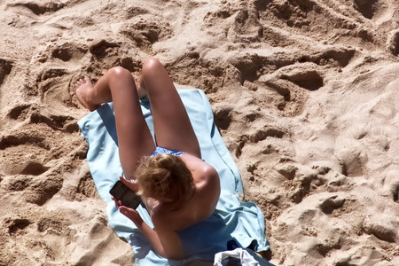 A girl in a blue swimsuit is lying on the sand and using her phone.の写真素材