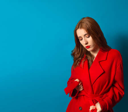 woman with make-up and red lips wearing her red trench coat on a blue background and with blank space.の写真素材