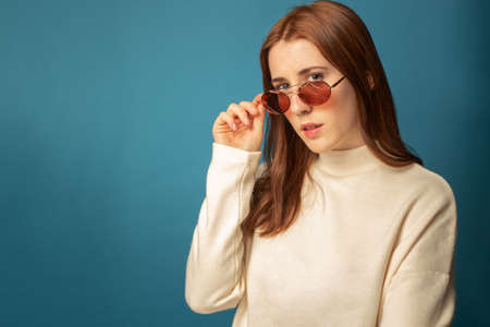 redhead woman with white sweater in a blue background looking over her orange glasses with a confident look.の写真素材