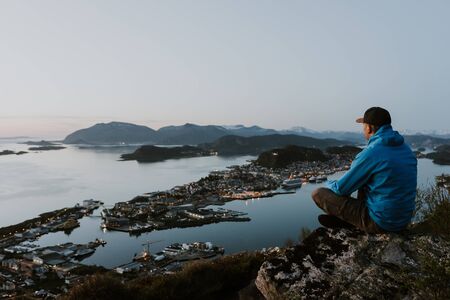 Blue and gray dressed traveller is looking at the norwegian city from the mountain. Travel and tourism concept. Shot during the blue hour.の写真素材