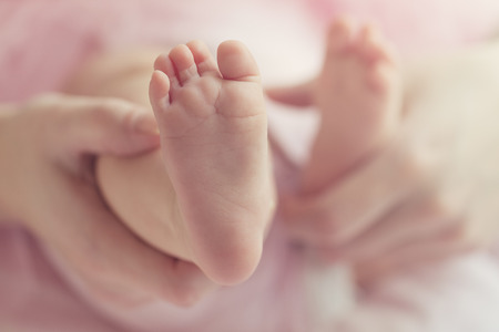 Closeup of mother hands holding cute tiny baby feet, showing baby foot. Toning. Pink Background. Horizontal.の写真素材