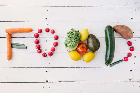 Flat lay of arranged raw vegetables in word Food on white wood.