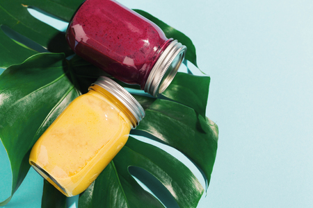 Tasty appetizing fresh made fruit smoothies in jars on monstera leaf on blue table. View from above with copy space. Tropical background.の写真素材
