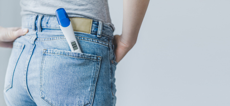Young woman or girl shows or holding positive pregnancy test with two stripes in jeans pocket. Lifestyle background. Pregnancy family concept. Horizontal.の写真素材
