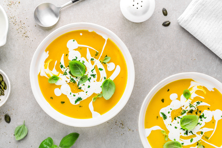 Tasty appetizing pumpkin vegetable creamy soup decorated with basil, cream and spices served in white bowls on grey table. View from above with copy space. Horizontal.  の写真素材