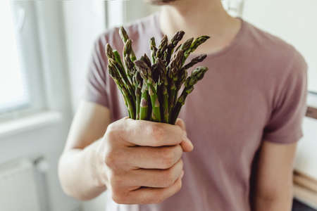 Clean eating or vegetarian concept. Young man holding fresh bundle of asparagus in the kitchen. Closeup.の写真素材