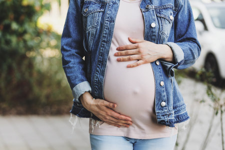 Pregnant woman in jeans jacket standing outdoors and touching belly.