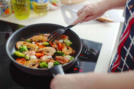 Woman staying at home kitchen and cooking shrimps with vegetables on pan. Home cooking or healthy cooking conceptの写真素材
