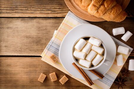 Cup of coffee with marshmallow, cinnamon sticks, brown sugar and croissant on wooden table. Top view.の写真素材