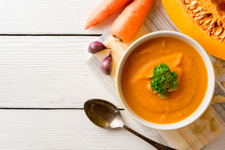 Pumpkin and carrot cream soup with pumpkin seeds and parsley in bowl on white wooden background. Top view with copy space.の写真素材