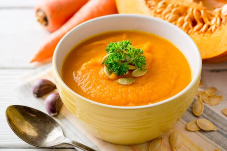 Pumpkin and carrot cream soup with pumpkin seeds and parsley in bowl on white wooden background. Selective focus.の写真素材