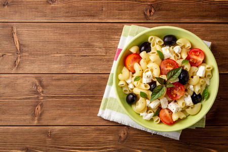 Pasta salad with cherry tomatoes, black olives, feta cheese and basil on wooden background. Top view. Copy space.の写真素材