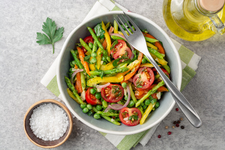 Salad with asparagus, bell pepper, cherry tomatoes, red onion and green peas in ceramic bowl on stone background. Top view.の写真素材