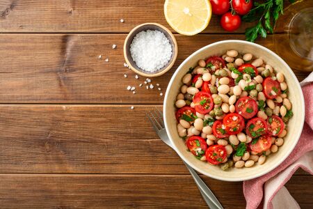 White bean salad with tomatoes, capers, garlic and parsley in bowl on wooden table. Top view. Copy space.の写真素材