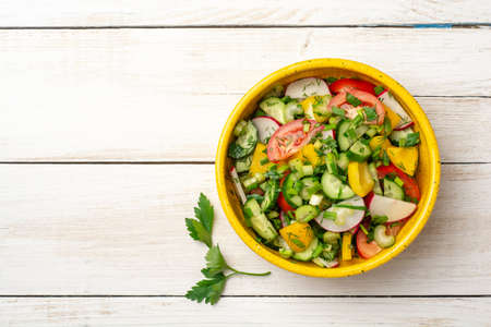 Fresh vegetables salad with radish, cucumber, bell pepper, tomato, celery and greens in ceramic bowl on white wooden rustic background. Top view, copy space.の写真素材
