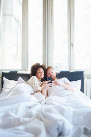 Young couple laughing together and using mobile phone while lying in bedの写真素材