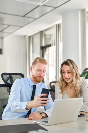 Two dedicated focused colleagues dressed in formal wear sitting in office and looking at smart phone. Start up business concept.の写真素材
