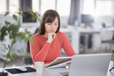 Charming Caucasian businesswoman sitting in moderna office and taking notes in notebook. In front of her laptop.の写真素材