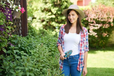 Beautiful Caucasian brunette with hat and dressed casual posing in her backyard with trowel in hand. Gardening time concept.の写真素材