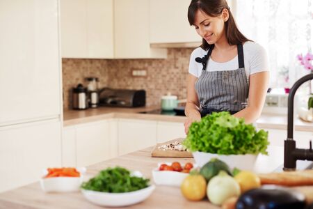 Beautiful smiling dedicated Caucasian brunette in apron standing in kitchen and chopping mushrooms. On table are lots of vegetables. Cooking at home concept.の写真素材