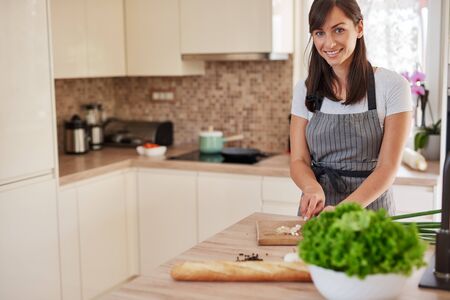Smiling Caucasian female chef in apron chopping leek in kitchen and preparing dinner. Preparation of domestic food concept.の写真素材