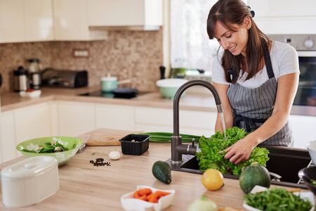 Cheerful attractive Caucasian woman in apron washing salad in kitchen. Dinner at home concept.の写真素材